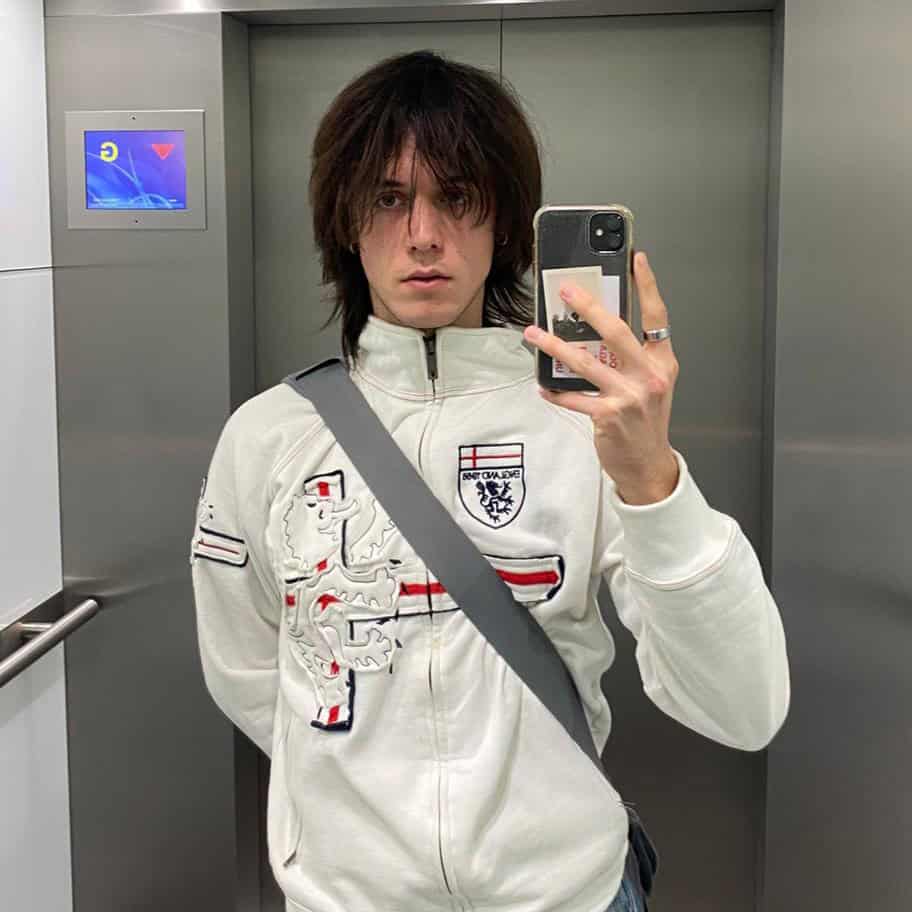A person with long dark hair takes a mirror selfie in an elevator, wearing a white England football jacket that proudly represents the team and a gray crossbody bag. Their phone partially covers their face.