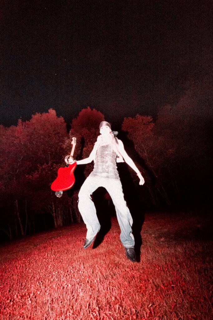 Piccolo wearing jeans and a tank top jumps in the air at night while holding a red electric guitar, with red lighting illuminating grass and trees in the background.