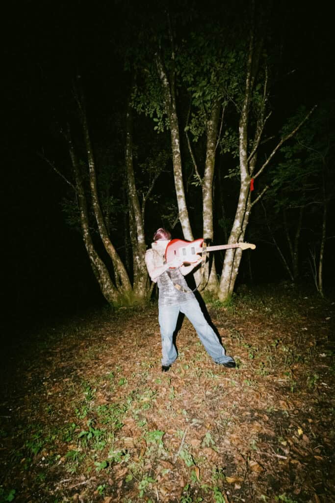 Piccolo stands outdoors at night in front of several trees, playing a red electric guitar passionately, with the ground covered in leaves and grass. The scene is illuminated by a bright, direct light.