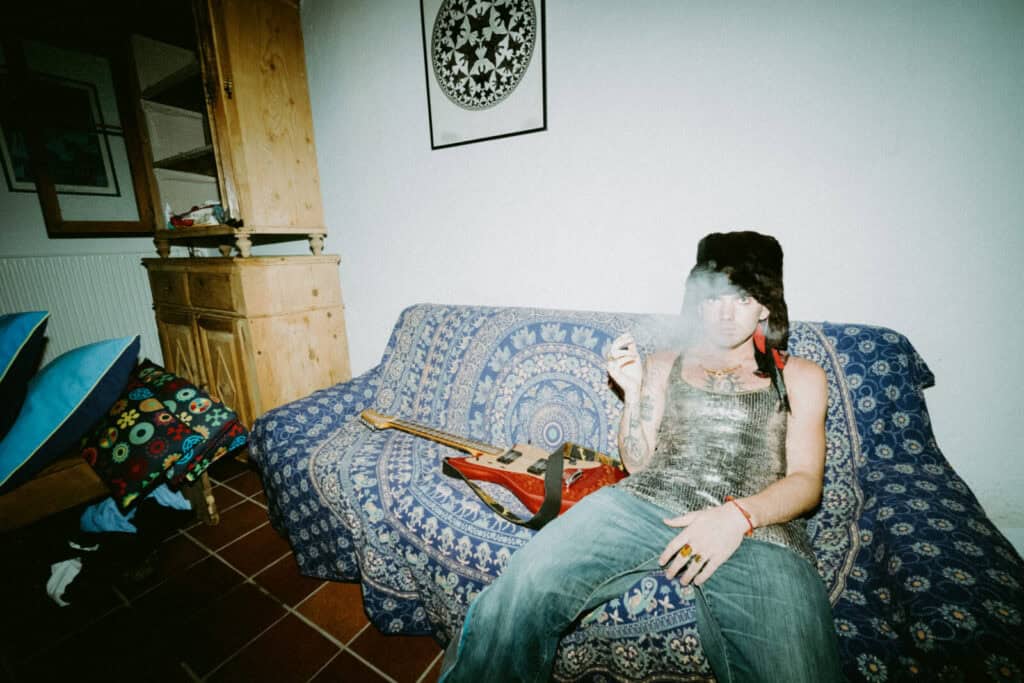 Piccolo in a furry hat and tank top sits on a patterned couch, exhaling smoke and holding a cigarette. An electric guitar rests beside them. The room has eclectic decor, with patterned pillows and artwork on the wall.