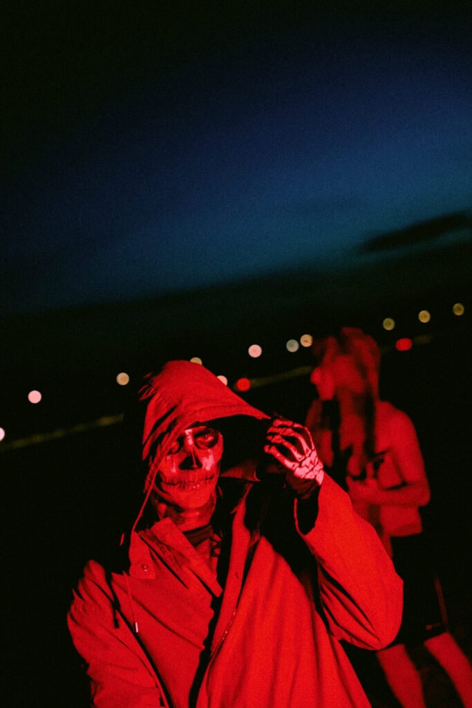 Piccolo wearing a red hooded coat and skull face paint stands outside at night, illuminated by red light, with blurred city lights and another person in the background.