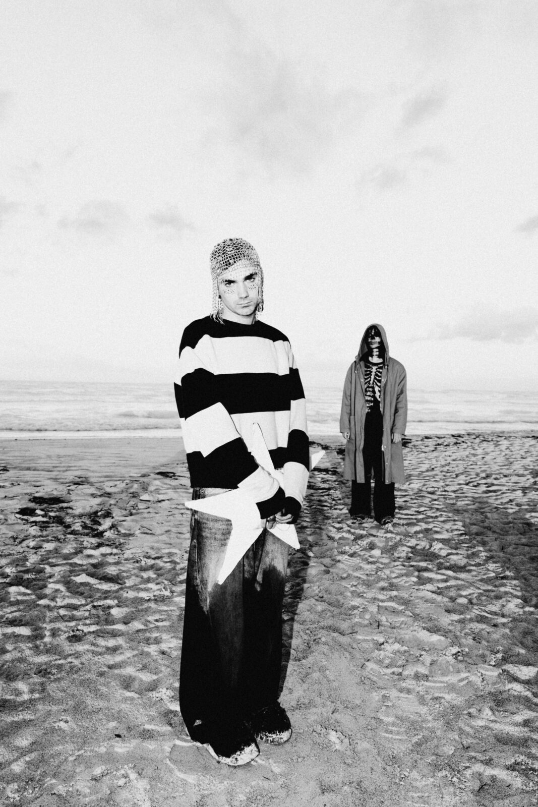 Piccolo and a second person stand on a beach in black-and-white. Piccolo wears a striped sweater and a chainmail-like headpiece. The second person, farther back, wears a long coat and sunglasses. The sky is cloudy.