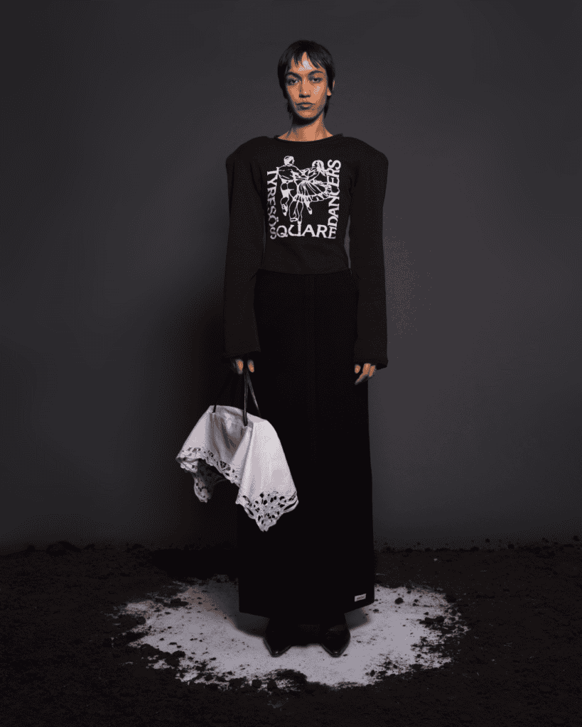 A person stands on a round patch of white powder against a dark background, wearing a black skirt, black graphic top with bold shoulders, and holding a folded white cloth bag with lace edging.