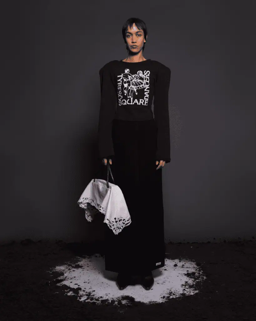 A person stands on a round patch of white powder against a dark background, wearing a black skirt, black graphic top with bold shoulders, and holding a folded white cloth bag with lace edging.