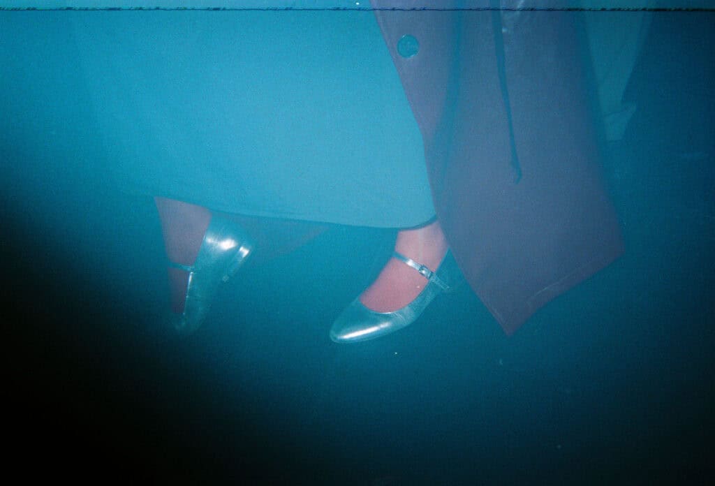 A person wearing shiny silver shoes and red tights stands on a dark floor, partially covered by a long coat—bathed in blue tint and black and white light leaks, evoking the mysterious vibe of a Lost Nightclub.