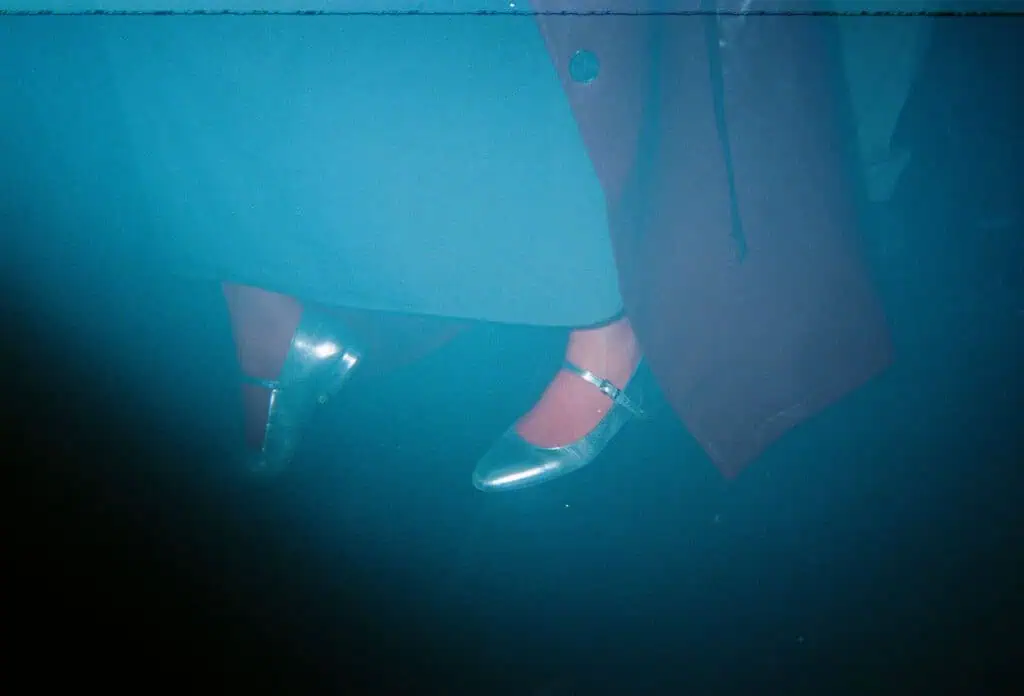 A person wearing shiny silver shoes and red tights stands on a dark floor, partially covered by a long coat—bathed in blue tint and black and white light leaks, evoking the mysterious vibe of a Lost Nightclub.