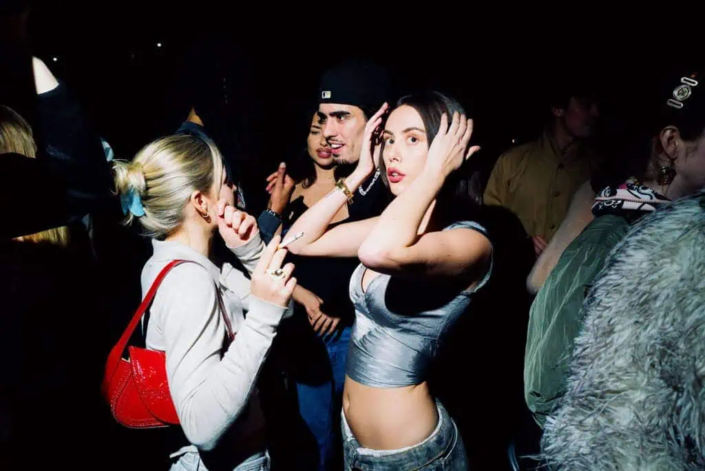 A group of young adults dance and socialize at Lost Club’s nighttime party. Two women in the foreground pose and gesture with their hands, while others mingle in the dimly lit background.