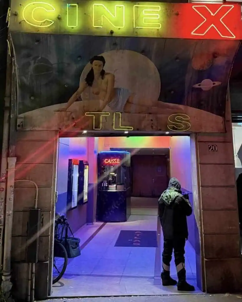A person in a hooded jacket stands at the entrance of a neon-lit cinema, its sign reading CINE X. With a vintage mural and colorful lights, the scene evokes the energy of a Lost Nightclub on a vibrant night.