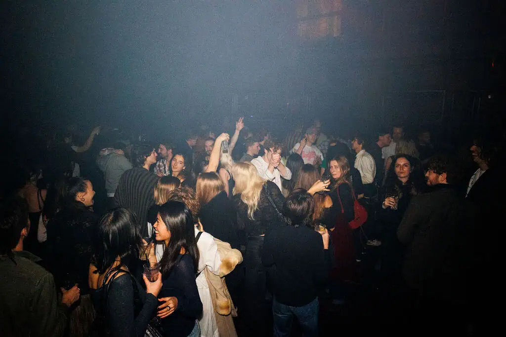A large group of people socialize and dance in the dimly lit Lost Nightclub, some holding drinks while others raise their hands in the air. The lively atmosphere pulses with energy and excitement.