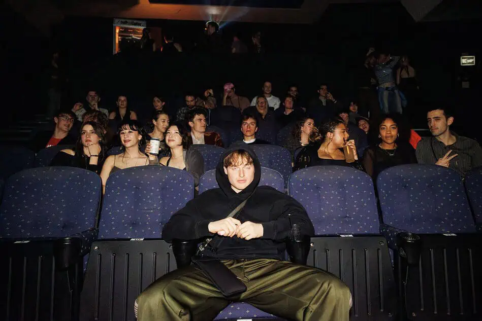 A young man in a black hoodie sits alone in the front row of a movie theater, facing the camera, while several people behind him talk and interact, evoking the isolating vibe of a lost nightclub.