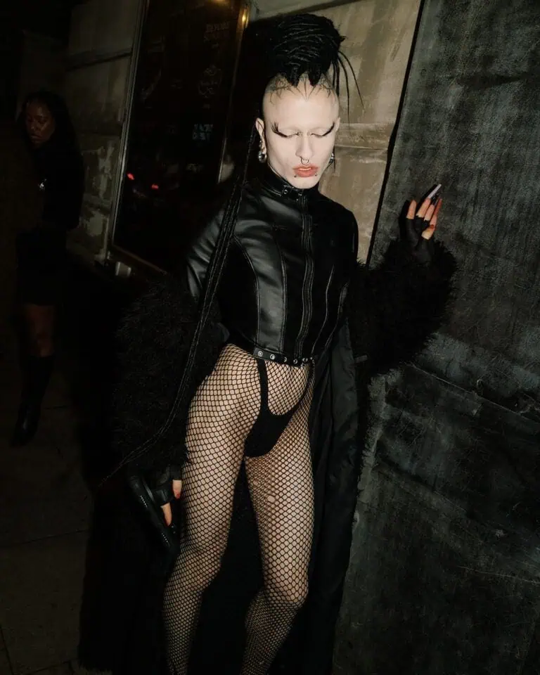 Person in dramatic goth-inspired outfit—black leather jacket, long textured coat, fishnet bodysuit, gloves, and bold makeup with high eyebrows and black lipstick—posing like a star outside the Lost Club against a city wall at night.