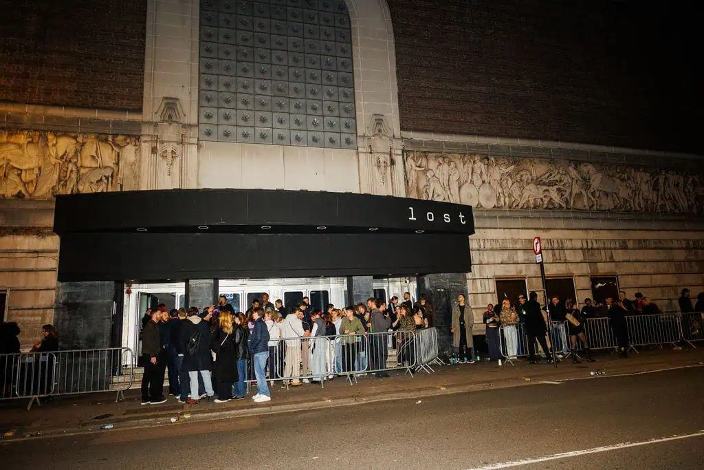 A crowd of people waits behind metal barricades outside Lost club, a building with a large black awning labeled "lost at night," featuring ornate stone carvings and tall windows.