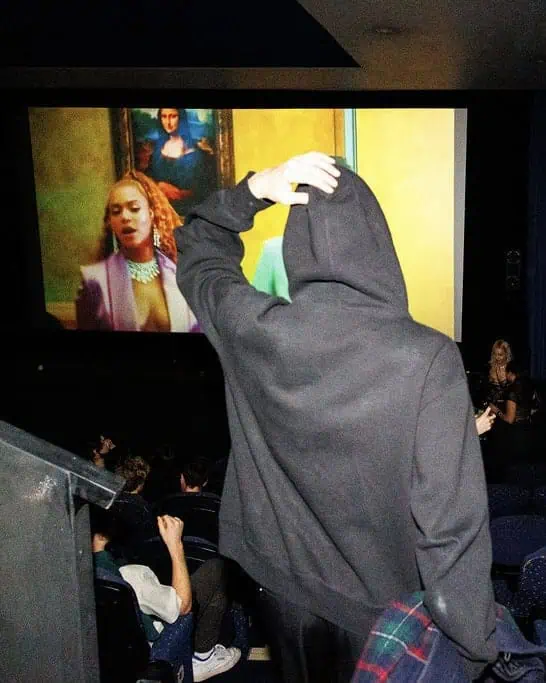 A person in a black hoodie stands facing a movie screen in a theater, scratching their head like they’ve entered the Lost Club. On the screen, a woman in a pink jacket and large necklace appears, with the Mona Lisa painting in the background.