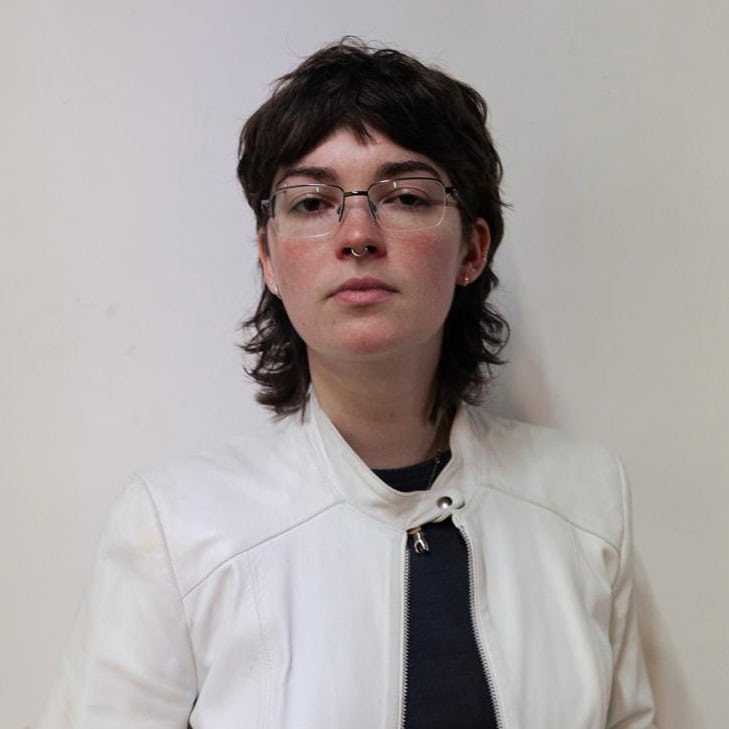 Lauren Bulla with short, dark, wavy hair and glasses wears a white jacket over a black shirt, standing against a plain light-colored wall and looking directly at the camera with a neutral expression.