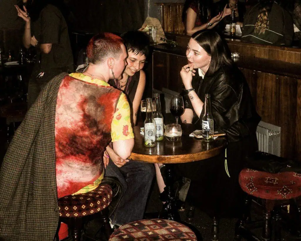 Three people sit closely around a small round table in a dimly lit bar, engaged in conversation and smiling. There are drinks and a candle on the table, and other patrons are visible in the background.