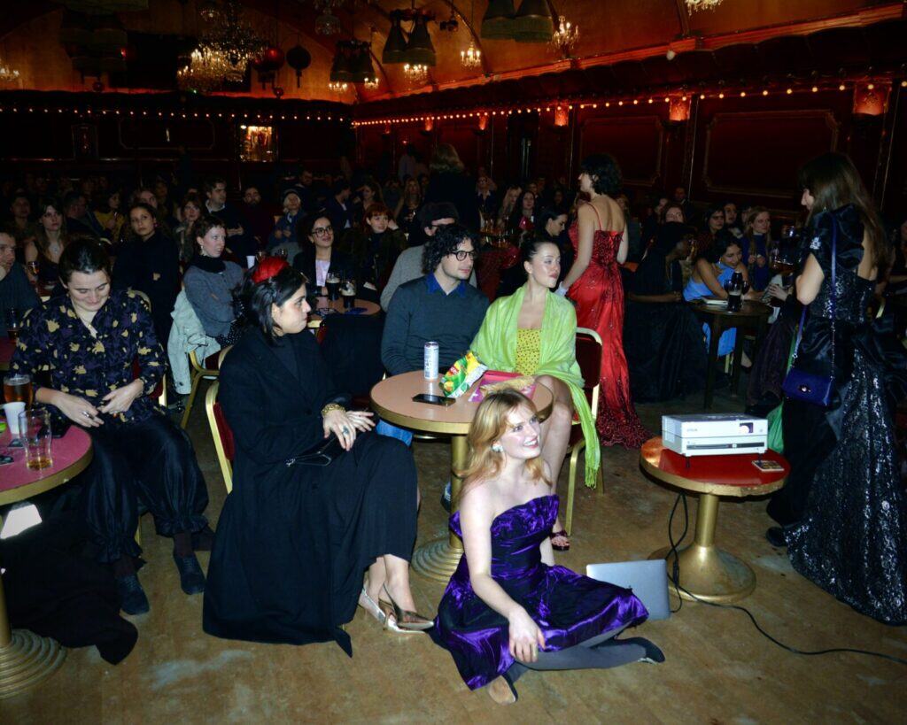 A crowded, dimly lit room with people seated at round tables, some dressed in formal or colorful attire. A woman in a purple dress sits on the floor near a small projector, while others watch something off-camera.