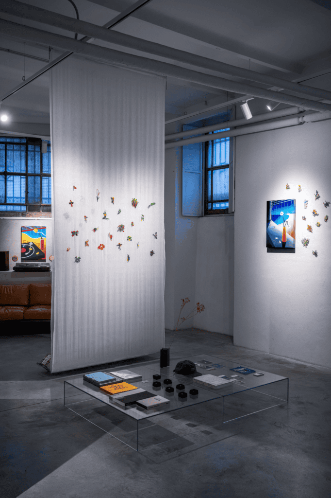 A modern art gallery interior inspired by Hiroki Yagi, featuring sheer white fabric hanging from the ceiling with colorful origami pieces. Artworks adorn white walls, while a glass coffee table with books sits on the concrete floor.