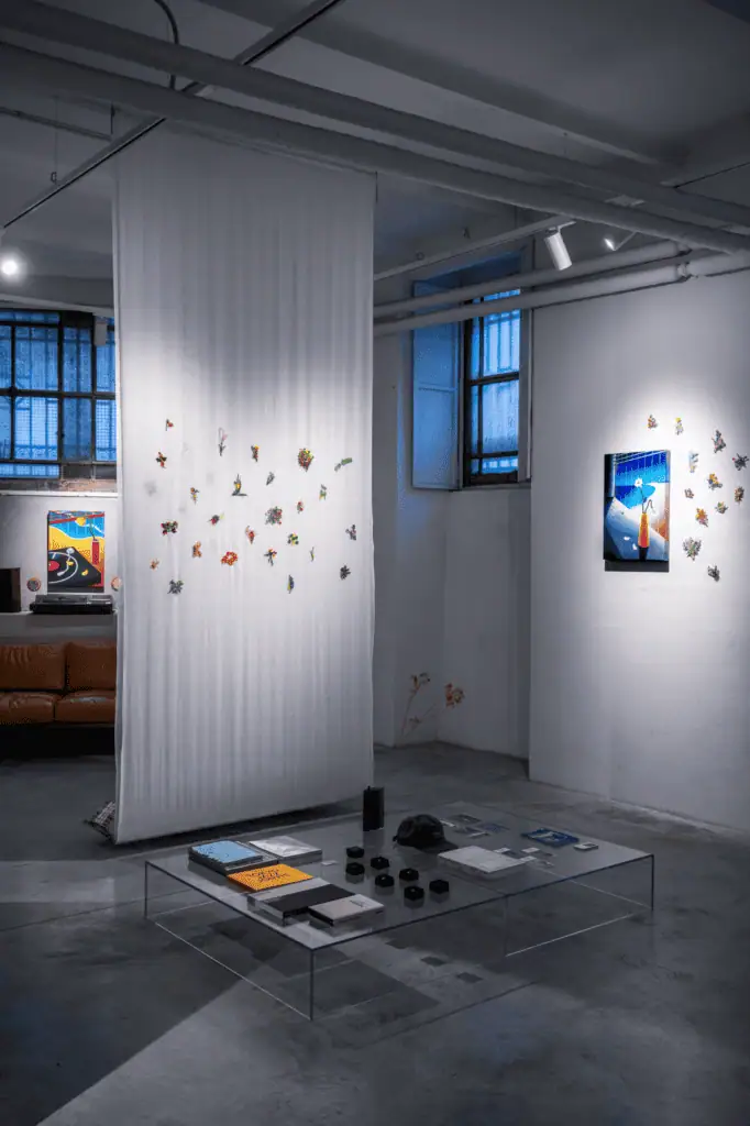 A modern art gallery interior inspired by Hiroki Yagi, featuring sheer white fabric hanging from the ceiling with colorful origami pieces. Artworks adorn white walls, while a glass coffee table with books sits on the concrete floor.