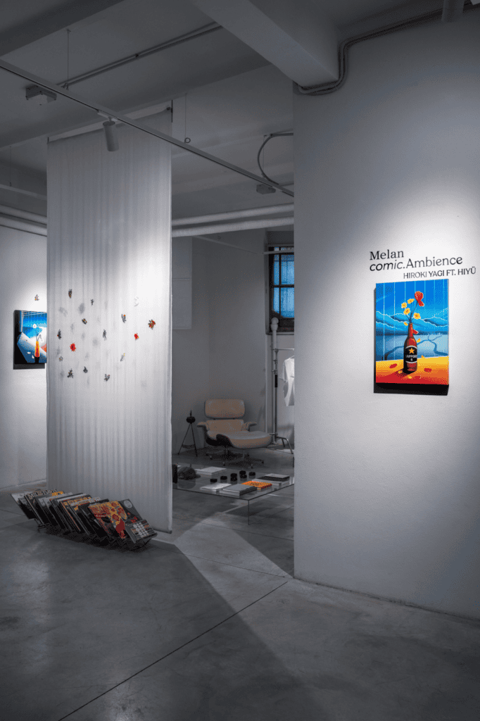 A modern art gallery with white walls displays colorful paintings, a hanging installation by Hiroki Yagi, and a collection of vinyl records stacked near the wall. A cozy seating area with a chair and coffee table is visible in the background.