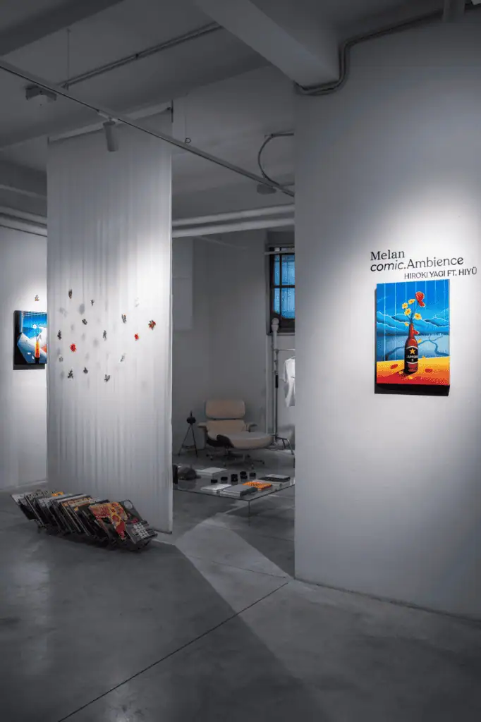 A modern art gallery with white walls displays colorful paintings, a hanging installation by Hiroki Yagi, and a collection of vinyl records stacked near the wall. A cozy seating area with a chair and coffee table is visible in the background.
