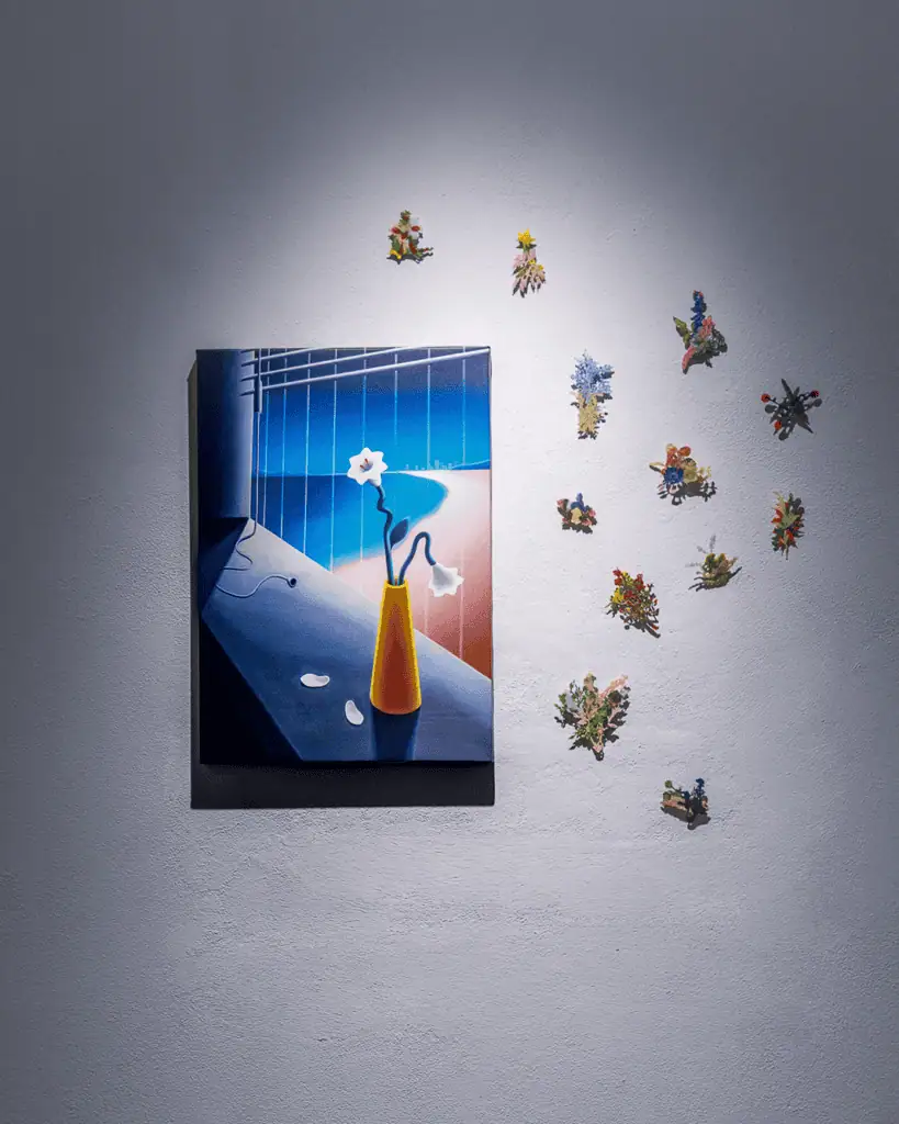 A painting by Hiroki Yagi of a yellow vase with white flowers sits on a dark table, hung on a light gray wall. Colorful, small, flower-shaped decorations are arranged on the wall to the right of the painting.