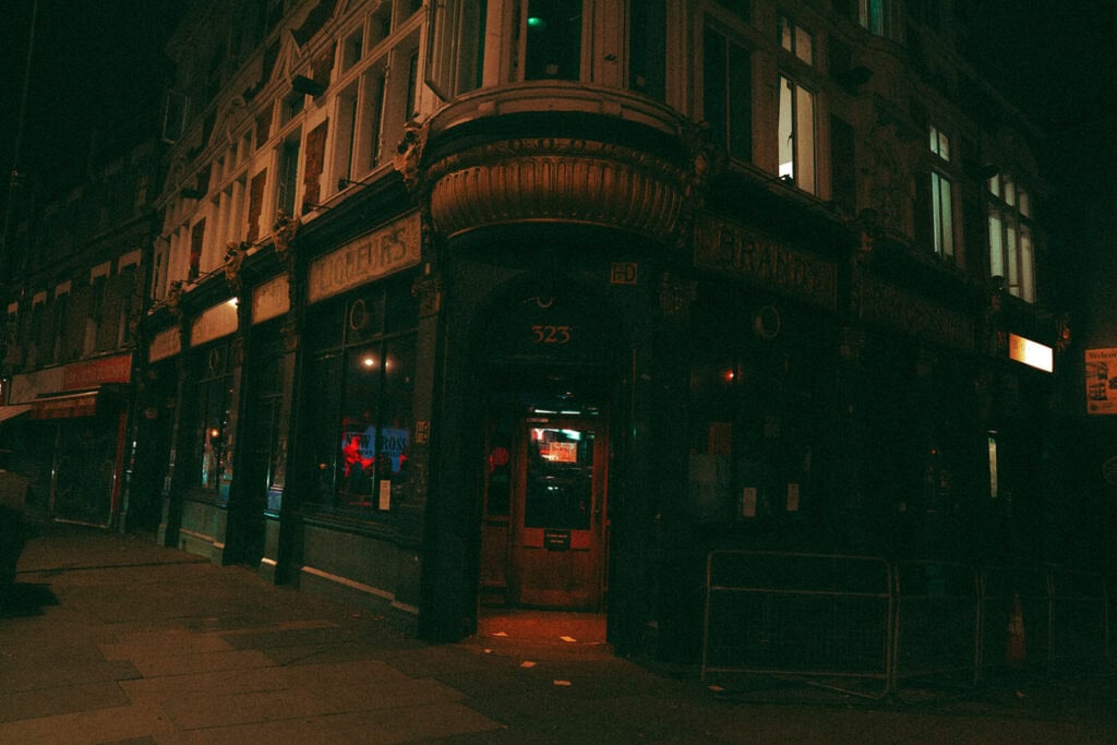 A dimly lit street corner at night features an old-fashioned pub with a large bay window, dark green facade, and glowing red lights inside. The entrance is marked with the number 323 above the door.
