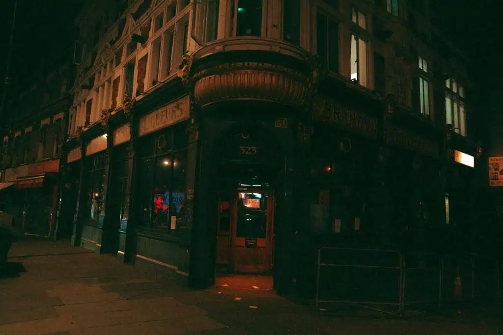 A dimly lit street corner at night features an old-fashioned pub with a large bay window, dark green facade, and glowing red lights inside. The entrance is marked with the number 323 above the door.