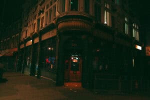 A dimly lit street corner at night features an old-fashioned pub with a large bay window, dark green facade, and glowing red lights inside. The entrance is marked with the number 323 above the door.