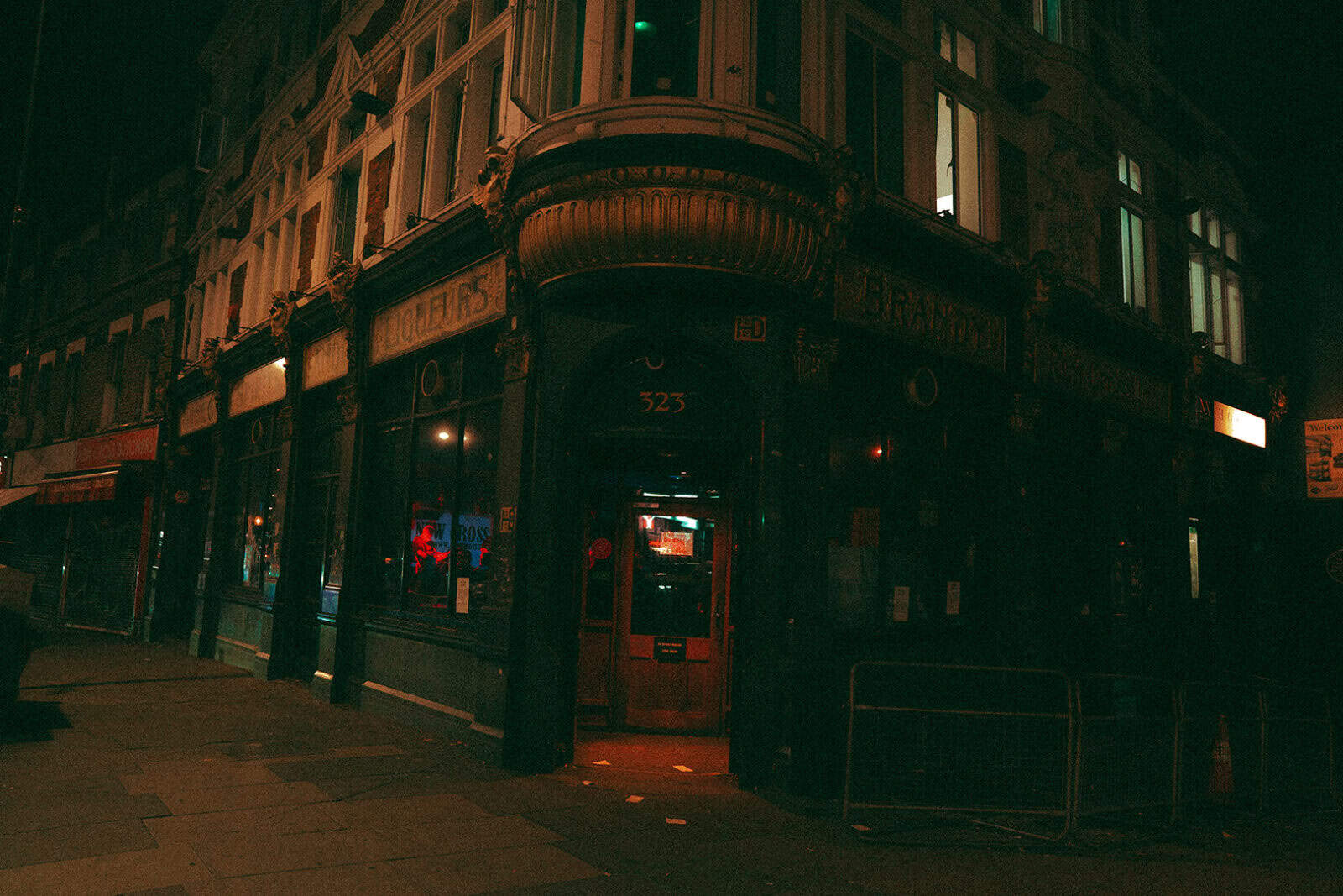 A dimly lit street corner at night features an old-fashioned pub with a large bay window, dark green facade, and glowing red lights inside. The entrance is marked with the number 323 above the door.