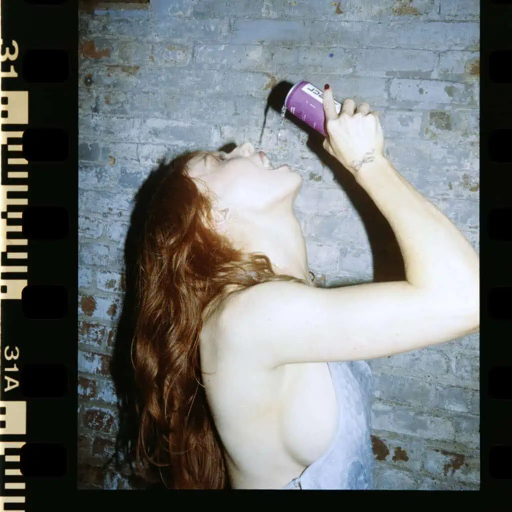 A person with long red hair tilts their head back and pours liquid into their mouth from a can, standing against a worn brick wall. The photo has a film strip border effect.