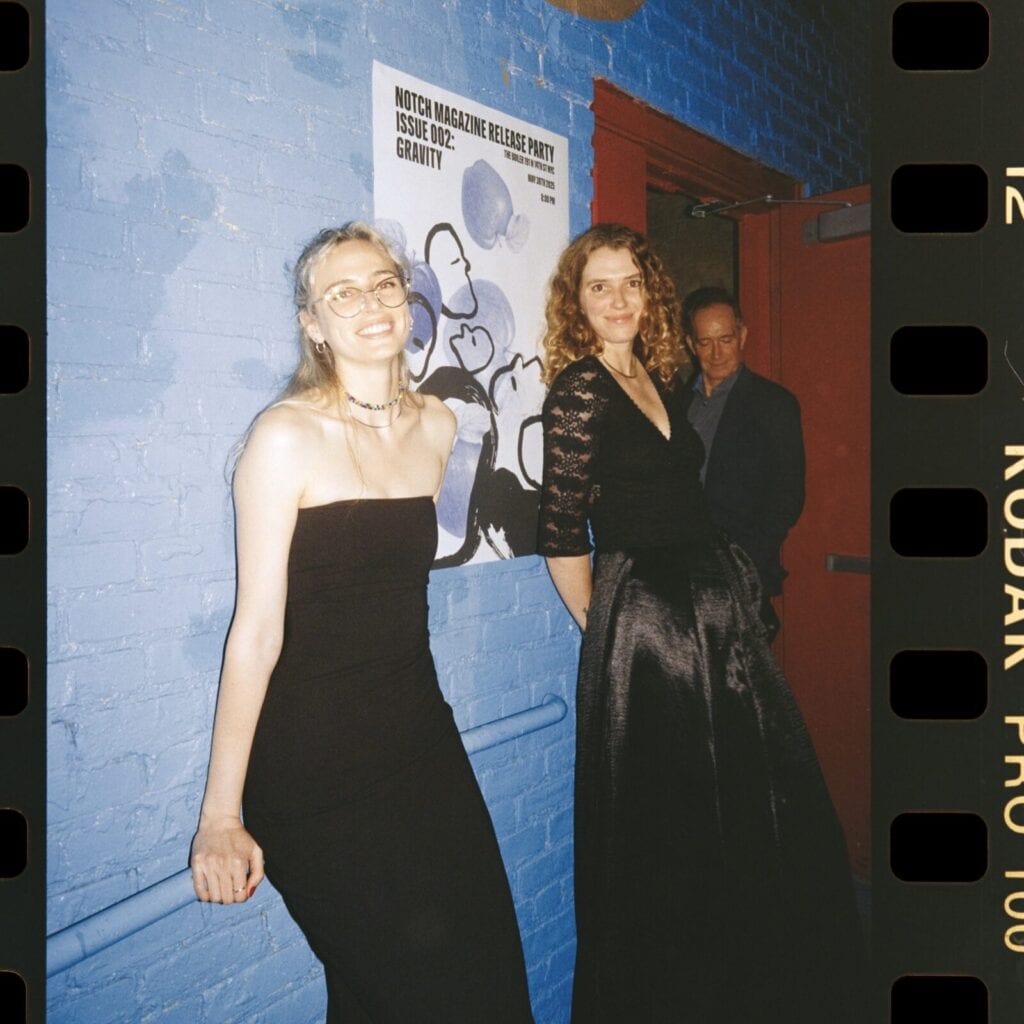 Two women in black evening gowns stand by a blue brick wall at an event, smiling. A man stands behind them near a red door. A poster for NOTCH MAGAZINE is visible on the wall. The image has a film photo style.