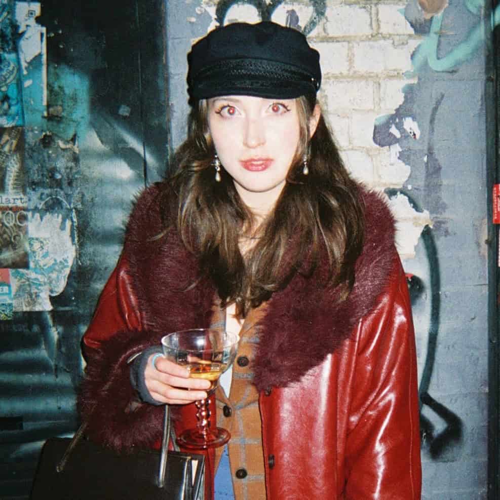 A woman with long brown hair, wearing a black cap and a red coat with a fur collar, holds a glass of wine and a brown bag. The team stands nearby as she poses in front of a graffiti-covered brick wall, looking at the camera.