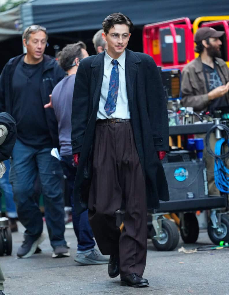 Timothée Chalamet in vintage clothing, including a long dark overcoat, pinstriped trousers, a tie, and glasses, walks on a film set surrounded by crew members and equipment.