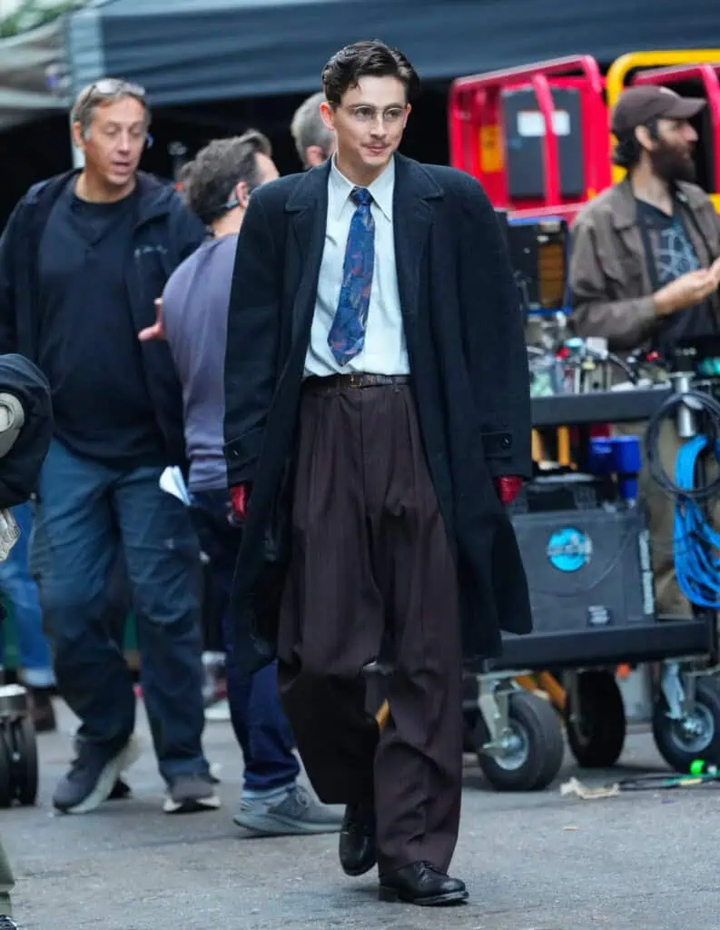 Timothée Chalamet in vintage clothing, including a long dark overcoat, pinstriped trousers, a tie, and glasses, walks on a film set surrounded by crew members and equipment.