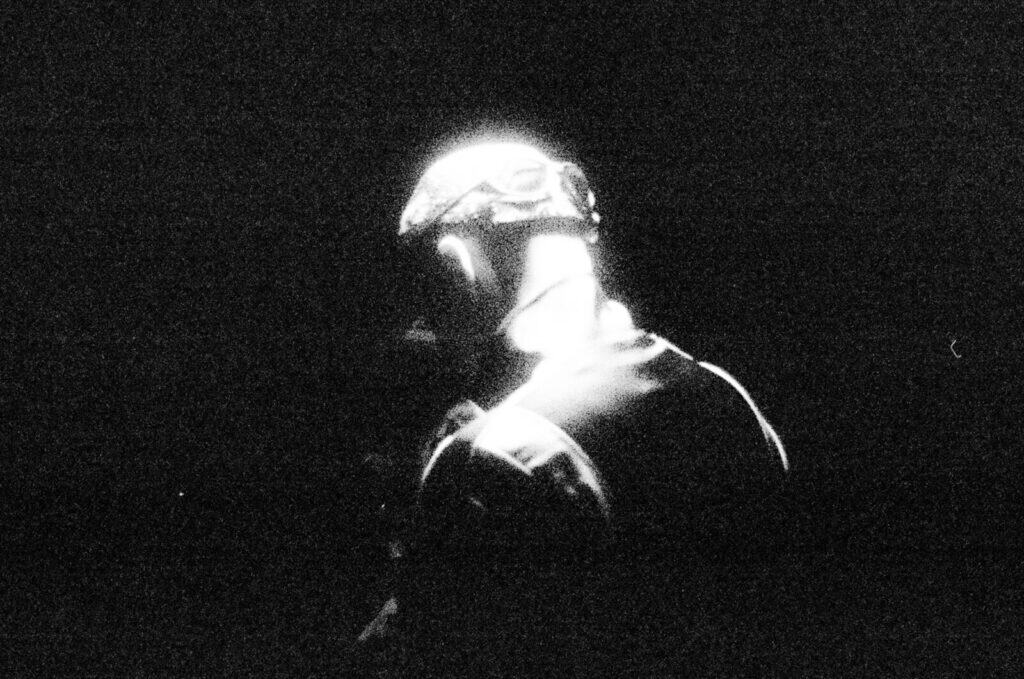 A person with their back turned, illuminated by bright light from above, stands against a dark, shadowy background, creating a dramatic and mysterious atmosphere.