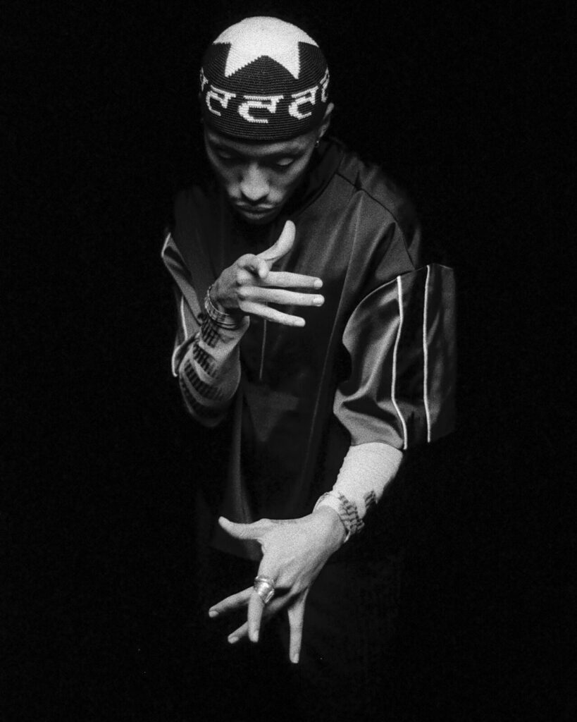 A person in a patterned hat and loose, dark clothing strikes an expressive pose against a black background, with their hands positioned artistically and head slightly bowed.