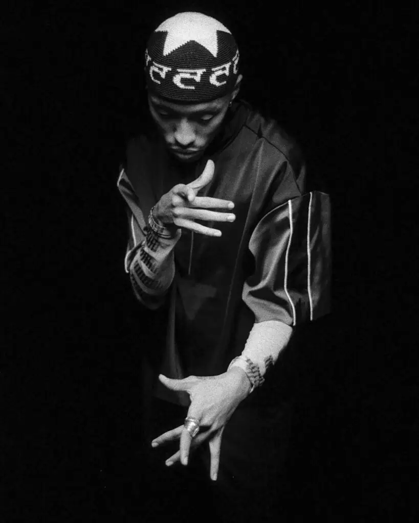 A person in a patterned hat and loose, dark clothing strikes an expressive pose against a black background, with their hands positioned artistically and head slightly bowed.