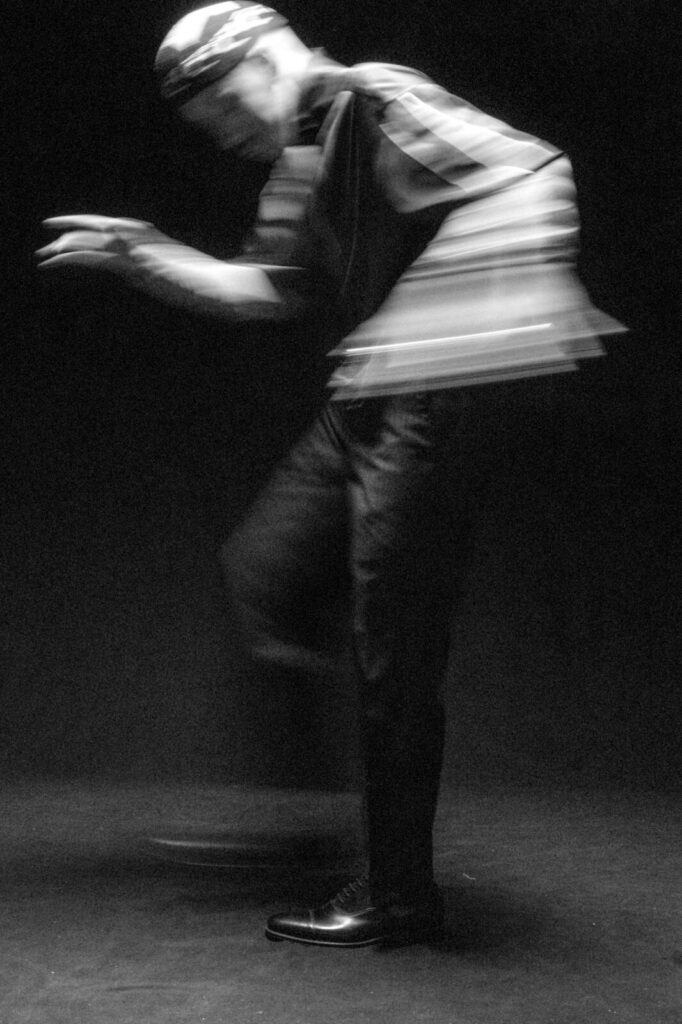 A black-and-white photo of a person in mid-motion against a dark background, creating a blurred effect. The person appears to be dancing or moving energetically, with one leg raised and arms outstretched.