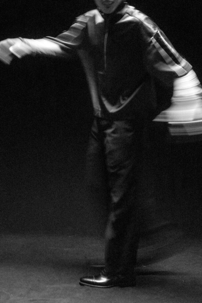 A black and white, blurry photo of a person in motion, wearing dark pants and a long-sleeve shirt, with arms outstretched and face partially visible, set against a dark background.