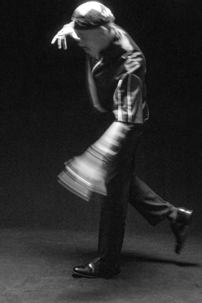 A person in motion, captured in black and white, wearing dark pants and a striped shirt, with one arm raised and one leg lifted, creating a blurred, dynamic effect against a dark background.