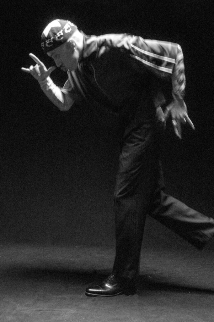 A person in a bandana, striped jacket, and dark pants strikes a dynamic dance pose, leaning forward with one leg bent, arms raised, and hands forming expressive gestures, set against a dark background.