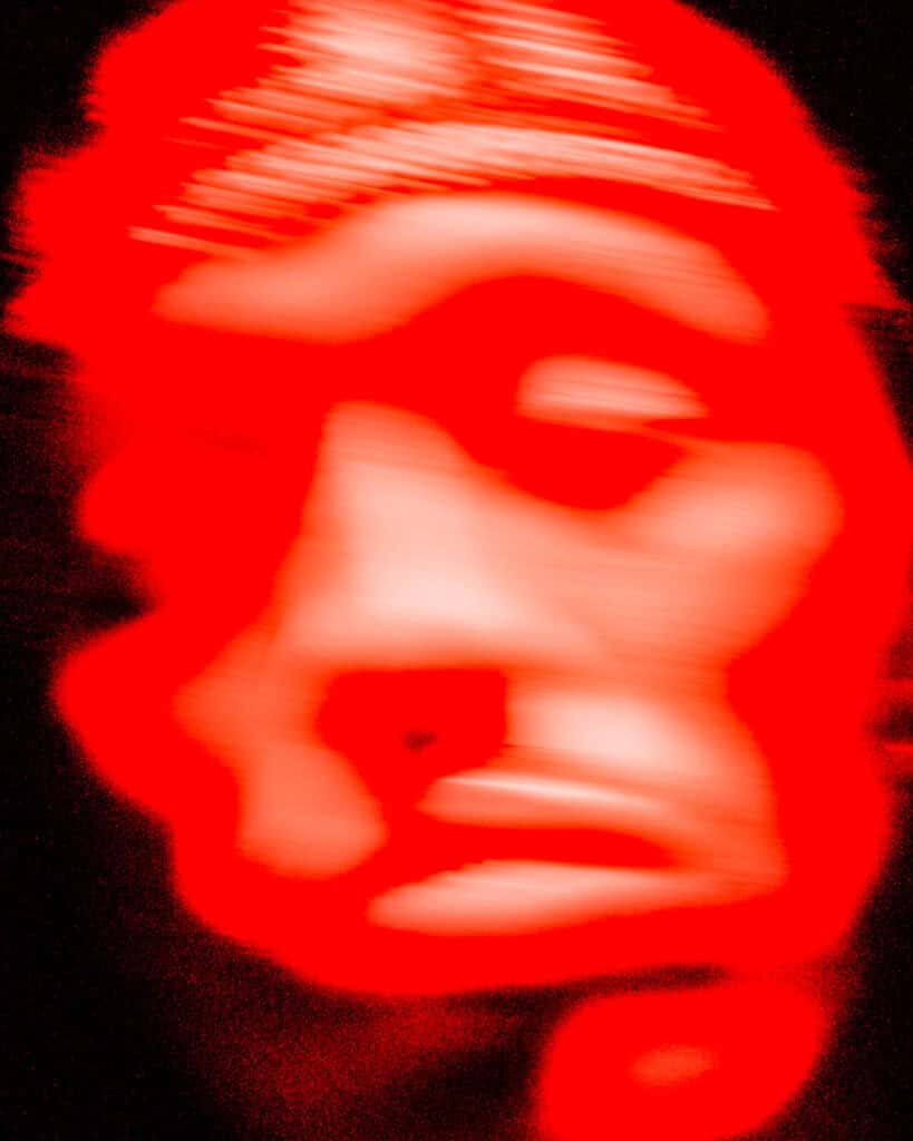 A heavily blurred, abstract red face set against a dark background, with distorted features and horizontal lines, creating an eerie and unsettling effect.