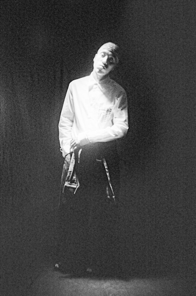 A person wearing a white shirt stands in a dimly lit, shadowy space, with their head tilted slightly and hands resting at their sides. The image has a grainy, black-and-white effect.