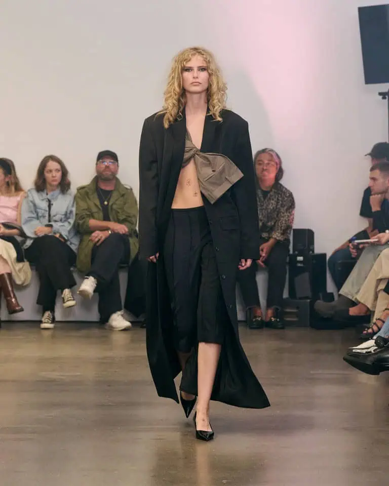 A model with curly blonde hair walks a runway wearing a long black coat, black skirt, black heels, and a large taupe bow top, in front of a seated audience in a minimalist indoor setting.