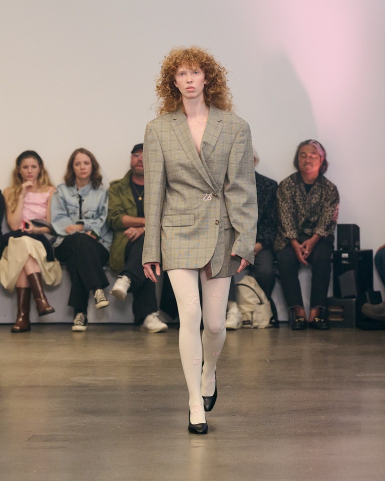 A model with curly hair walks a runway wearing an oversized gray plaid blazer, white tights, and black shoes. Seated audience members watch in the background against a plain wall.