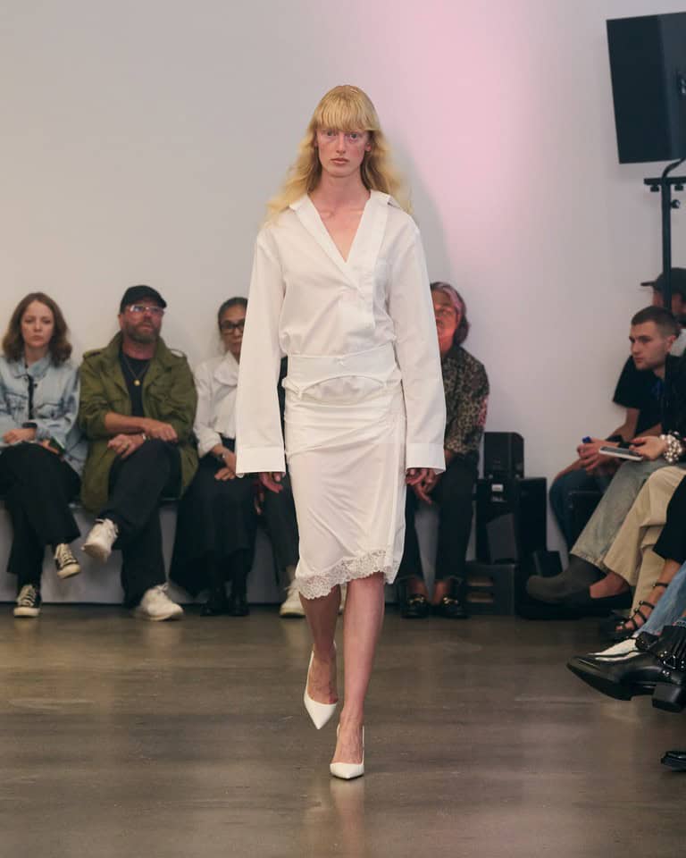 A model with long blonde hair walks a runway wearing a white blouse with wide sleeves and a matching white skirt with lace trim, while audience members sit and watch from both sides.