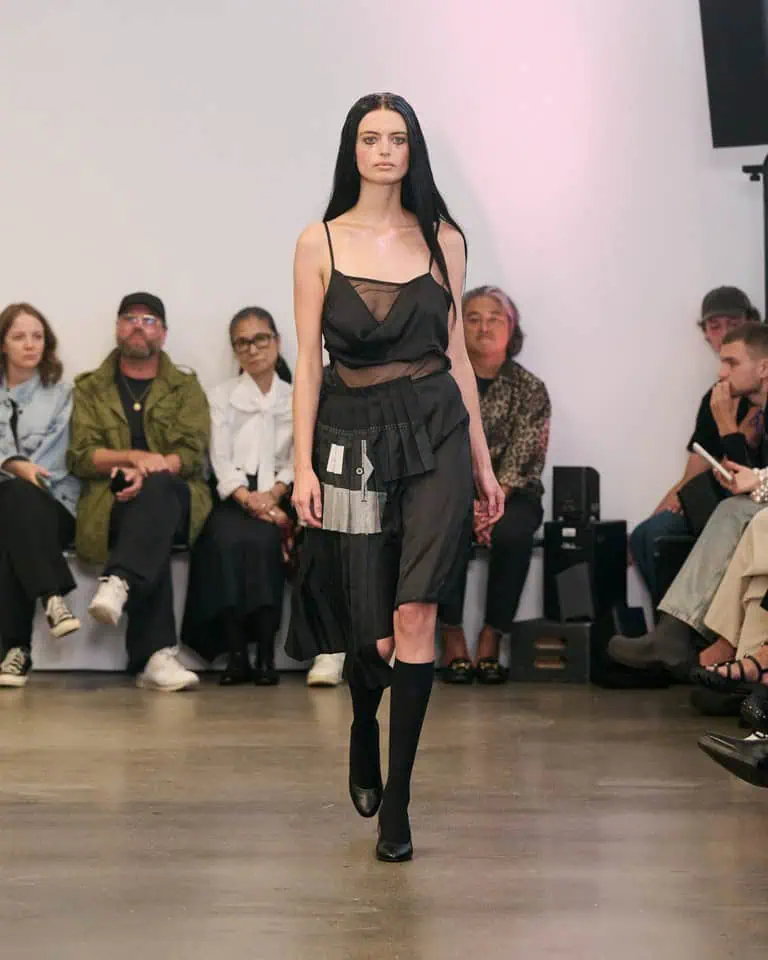 A model with long dark hair walks a runway wearing a sheer black dress with layered fabric details and knee-high black socks. Seated audience members watch from both sides of the runway in a minimalist room.