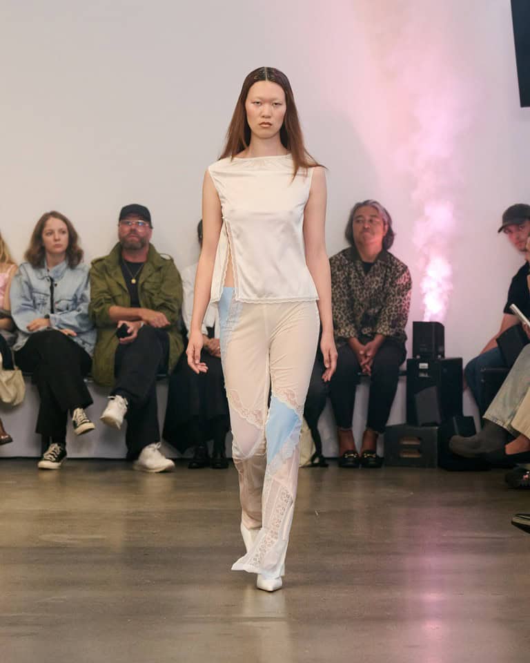 A model walks a runway wearing a sleeveless, off-white satin top and sheer pants with light blue and white lace panels. Several seated spectators watch in the background.