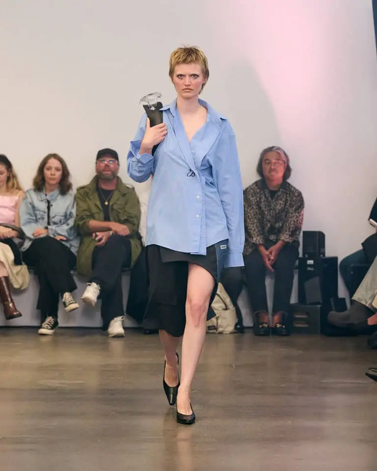 A model walks a runway wearing an oversized blue shirt, black skirt, and black heels, holding a large crystal-like object on their shoulder. Seated audience members watch in the background.