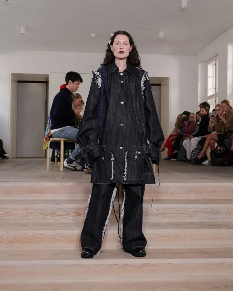 A model stands on wooden steps wearing an oversized black denim jacket and matching wide-legged pants with white stitching, in front of a seated audience at a fashion show in a minimalist indoor space.